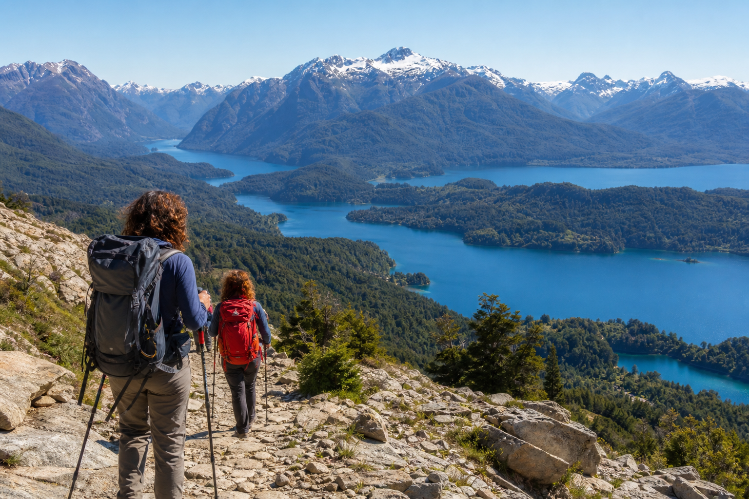Senderistas caminando por un sendero de montaña en Bariloche con lagos de color azul, bosques y picos nevados al fondo.